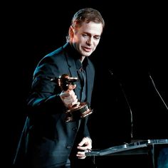 a man standing at a podium holding an award in front of a microphone and wearing a suit