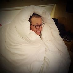 a man with glasses is wrapped in a blanket and smiling at the camera while lying in bed