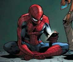 a spider - man sitting on the ground next to a person holding a cell phone