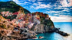 the village is perched on top of a cliff by the water's edge, with blue skies and clouds in the background