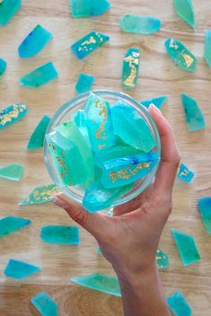 a hand holding a glass bowl filled with blue and green seaglass pieces on top of a wooden table