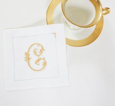 a white napkin with a gold monogram on it next to a coffee cup and saucer