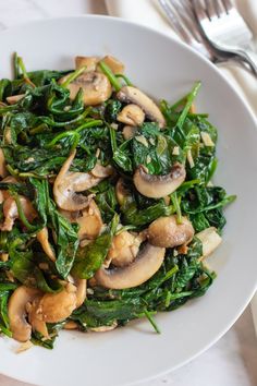 spinach and mushrooms in a white bowl on a plate with silverware next to it