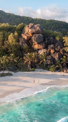 two people are walking on the beach next to some rocks and trees in the background