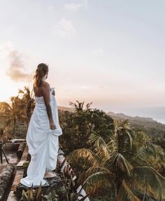 a woman in a white dress standing on a wooden platform overlooking the ocean and palm trees