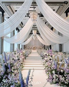 an aisle decorated with white drapes and purple flowers on either side is a chandelier that hangs from the ceiling