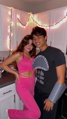 a man and woman standing in a kitchen with lights on the wall behind them, posing for a photo