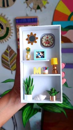 a person holding up a miniature shelf with plants and pictures on the wall behind it