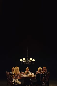 four women sitting at a table eating food in front of a chandelier on a dark background