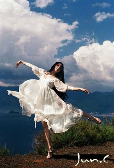 a woman in a white dress is dancing by the water with her arms spread out