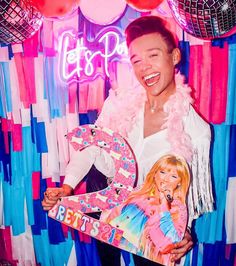 a man holding a barbie doll in front of a party backdrop with balloons and streamers