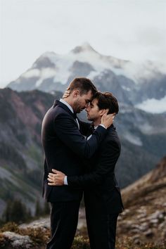 two men in suits embracing each other on top of a mountain with snow capped mountains behind them