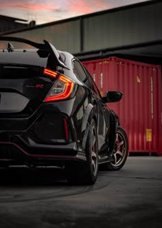 the rear end of a black car parked in front of a shipping container at dusk