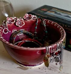 a red bowl filled with lots of jewelry next to a wine glass and box on top of a wooden table