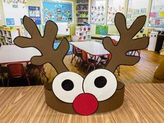 a cardboard reindeer head sitting on top of a table in front of a classroom filled with desks