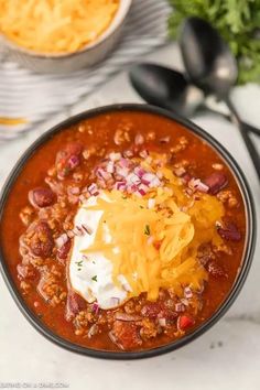 a bowl filled with chili and cheese on top of a white countertop next to silverware