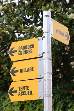 three yellow signs pointing in different directions on a metal pole next to trees and bushes