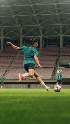 a woman kicking a soccer ball on top of a field in front of an empty stadium