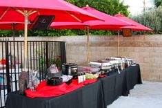 an outdoor buffet table with red umbrellas and food items on it, in front of a fence