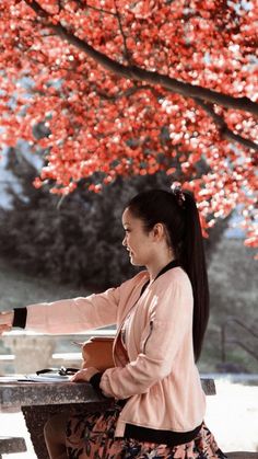 a woman sitting at a table under a tree with red flowers on it's branches