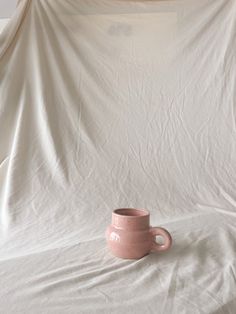 a pink cup sitting on top of a white sheet covered tablecloth in front of a wall