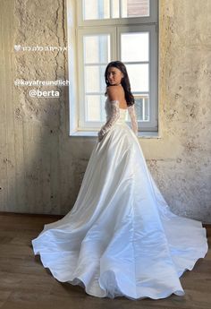 a woman in a white wedding dress standing by a window with her back turned to the camera