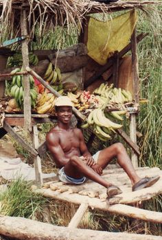 a man sitting in front of a bunch of bananas on top of a wooden bench