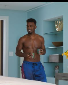 a shirtless man holding a cup in his hand while standing next to a bed