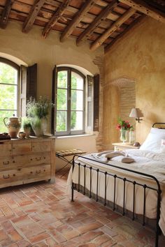 a bed sitting in a bedroom next to two windows and a table with flowers on it