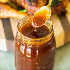 a spoon full of bbq sauce sitting on top of a wooden cutting board with chicken wings in the background