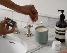 a person lighting a candle in front of a sink with soap and lotion on it