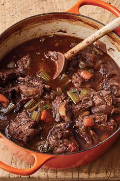 a pot filled with beef and carrots on top of a wooden table next to a spoon