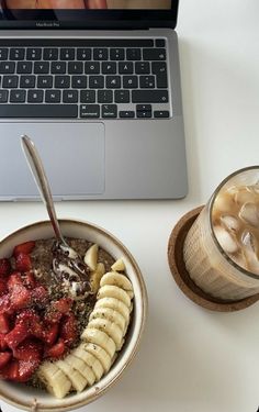 a bowl of fruit next to a laptop on a white table with a cup of coffee