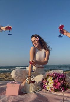 a woman sitting on the beach with two wine glasses in her hand and a cake