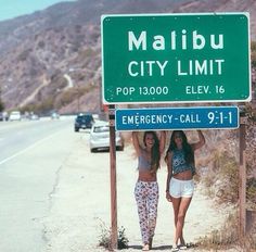 two women standing under a street sign on the side of the road with mountains in the background