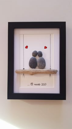 two pebbles sitting on a swing with hearts hanging from the strings, framed in a black frame