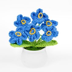 blue crocheted flowers in a white vase on a white surface with green leaves