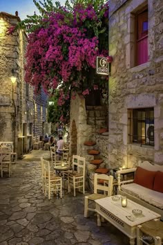 an alley way with tables and chairs on both sides, purple flowers growing over the wall