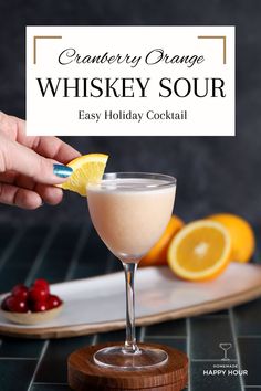 a hand holding a lemon wedge over a glass filled with an orange and cranberry orange whiskey sour