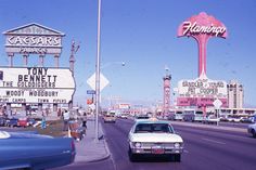 an old car driving down the road in front of some neon signs and other buildings