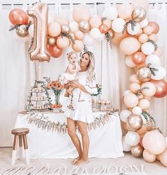 a woman holding a baby standing in front of a cake table with balloons and garlands