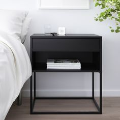 a black night stand with magazines on it next to a white bed and potted plant