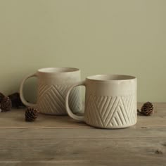 two coffee mugs sitting next to each other on top of a wooden table with pine cones