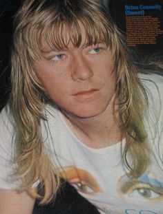 a man with long blonde hair wearing a white t - shirt and looking at the camera