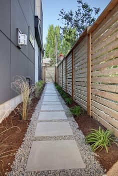 a stone path between two buildings in front of a wooden fence and planter area