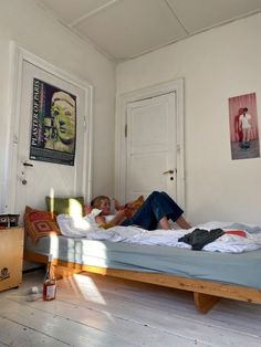 two people laying on a bed in a room with white walls and wooden floors,