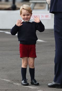 a little boy in red shorts and black sweater standing next to a man with his hand up