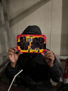 a person wearing a hood and holding up a box with graffiti written on it in front of their face