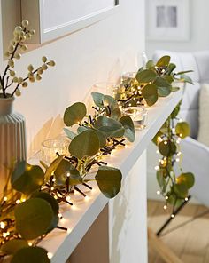 a mantle with candles and greenery on it in front of a white wall decorated with lights