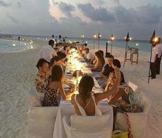 a group of people sitting at a long table on the beach eating food and drinking